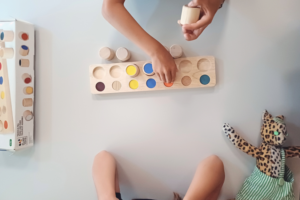 Blick von oben auf ein Holzspiel auf einem Tisch, umgeben von zwei Paar Kinderarmen. Die oberen Arme setzen eine Spielfigur aufs Brett, unten rechts liegt ein Stofftier-Leopard.