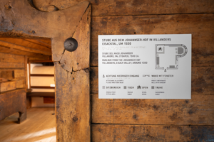 Zugang zur Stube aus dem Johannser-Hof in Villanders ©Wolfgang Lackner. Bildbeschreibung: rechts ein Schild an der Wand mit haptisch lesbarer Raumbeschreibung inklusive Grundriss, links ein kleiner Einblick durch die Türöffnung in die Stube. 