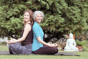 Bild: Die Kursleiterinnen Daniela Maltan und Lea Stoll sitzen Rücken an Rücken im Schneidersitz auf einer Yoga-Matte mit Blick in die Kamera, rechts neben ihnen steht eine kleine Buddha-Statue auf der Wiese.