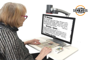 Man sieht eine ältere Dame am Schreibtisch sitzen. Mit ihren Händen berührt sie eine vor ihr auf den Tisch liegende Zeitung; sie blickt auf den Bildschirm des Geräts 