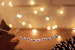 Auf einer Schnur sind Buchstaben-Würfel aufgefädelt, die nebeneinander die Worte „FROHE WEIHNACHTEN“ bilden. Dahinter sind verschwommen stimmungsvolle Lichtpunkte zu sehen; am linken und rechten Bildrand sind Ausschnitte eines Tannenzapfens und eines kleinen Deko-Weihnachtsbaums zu sehen.