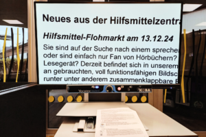 Bild 1: Man sieht ein Bildschirmlesegerät mit großem Bildschirm und dem lesbaren, stark vergrößerten Text „Neues aus der Hilfsmittelzentrale: Hilfsmittel-Flohmarkt am 13.12.24“ und mehr.