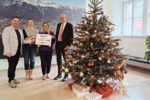 Andreas Michel (Vorsitzender Hauptgruppe II der younion), Birgit Brugger (Co-Leiterin der BSVT-Frühförderung), Sabine Karrer (Obfrau des BSVT), die ein symbolisches Spenden-Schild in Höhe von € 1.500,- in die Kamera hält, und DI Helmuth Müller (Vorsitzender des Vorstandes der IKB) (v.l.n.r.); neben ihnen ein hübsch geschmückter Christbaum und im Hintergrund ein großes Bild von Innsbruck mit der Nordkette im Hintergrund.
