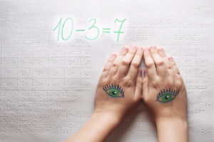 Bildbeschreibung: Zwei Kinderhände, die Braille-Schrift lesen, sind auf der Handrückseite mit Augen bemalt. Oben links leuchtet die Mathematikaufgabe 