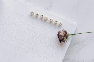Ein Blatt mit Brailleschrift liegt auf einer Marmorplatte. Darauf Buchstabenwürfel, die „Braille“ ergeben. Eine getrocknete, rosafarbene Rose ohne Dornen liegt quer darüber – ein stilles, poetisches Dankeschön an die Blindenschrift.