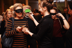 Besucherinnen und Besucher mit Augenbinden werden von einer Begleitperson zu ihrem Platz geführt; Foto: Max Wegscheidler.