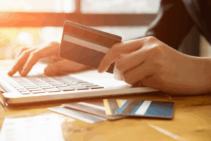 Eine Person tätigt eine Überweisung am Laptop und hält dabei eine Kreditkarte. Weitere Karten liegen auf dem Tisch.